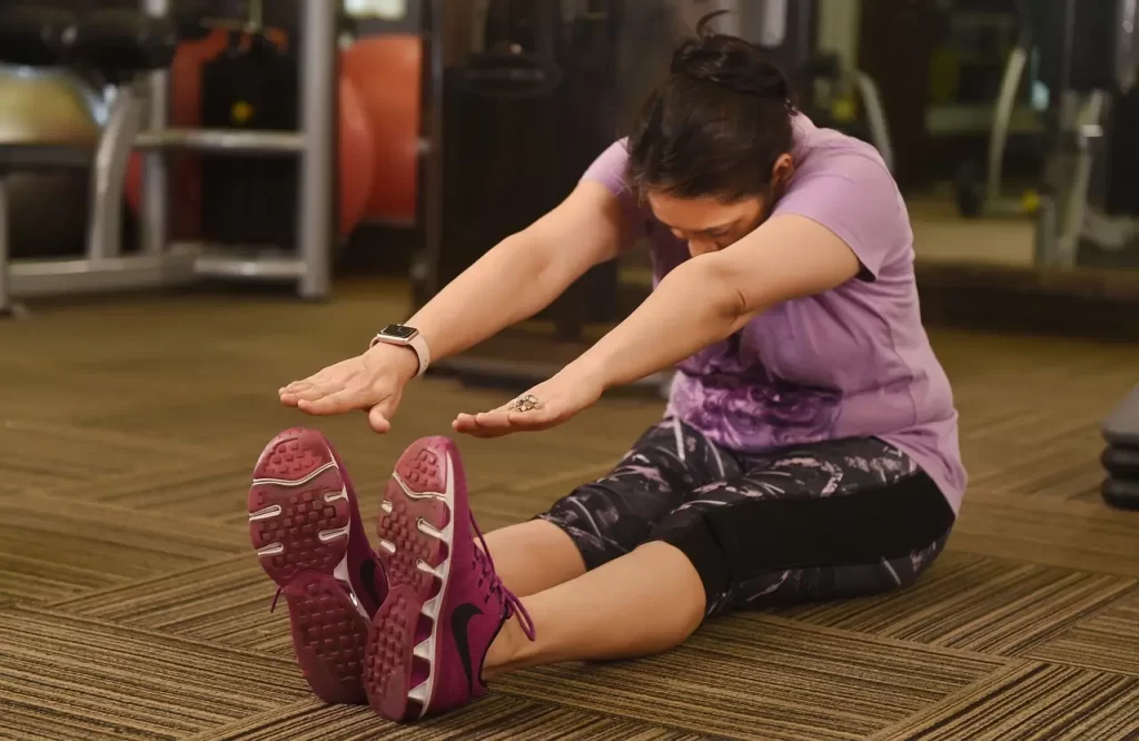 A person kneeling on the floor, wearing casual clothing and shoes, with their elbows resting on their knees. The environment is indoors with visible flooring. - Exercise Is Keeping People Fat But No One Wants to Talk About It