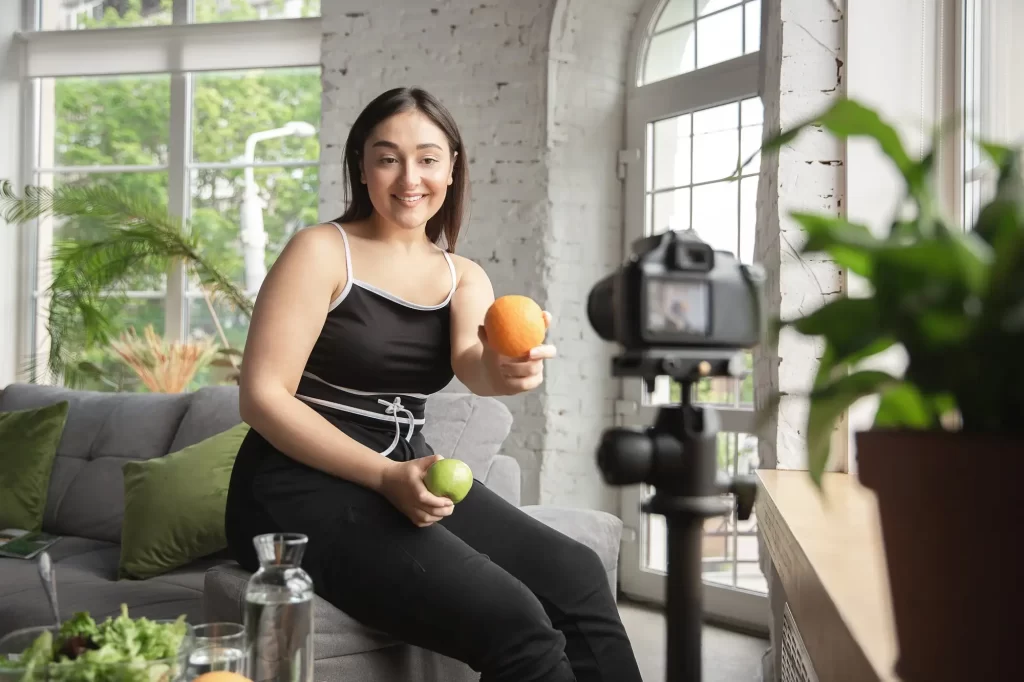 A woman sitting on a couch, smiling while holding an orange, surrounded by houseplants and a vase with flowers, with sunlight coming through a window. -Exercise Is Keeping People Fat But No One Wants to Talk About It