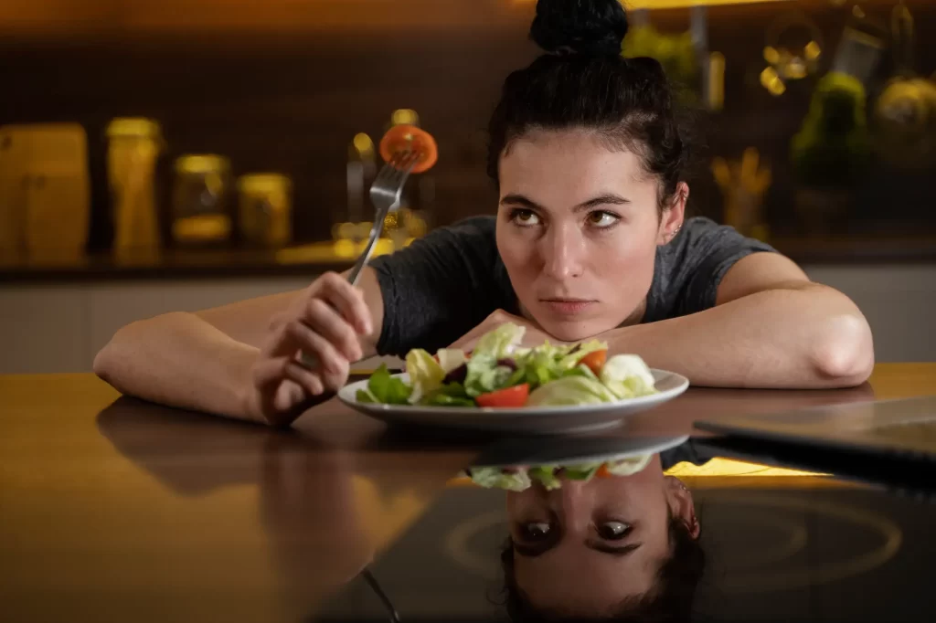 Exercise Is Keeping People Fat But No One Wants to Talk About It woman eating a salad in a warmly lit kitchen.