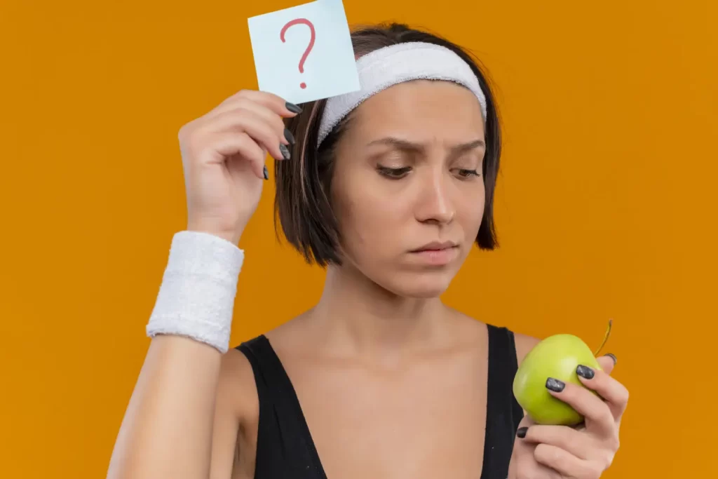 Exercise Is Keeping People Fat But No One Wants to Talk About It - A young fitness woman in sportswear holding a note with a question mark and a green apple, reflecting on the debate about whether