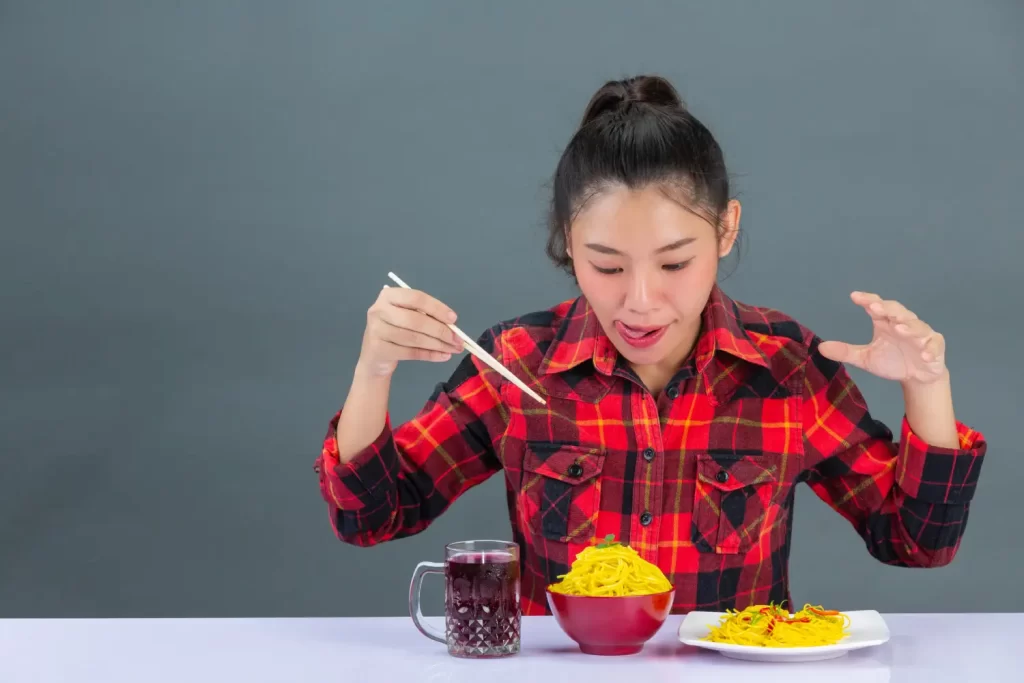 A young woman in a plaid shirt enthusiastically eating spaghetti with chopsticks, enjoying her meal at home. - Exercise Is Keeping People Fat But No One Wants to Talk About It