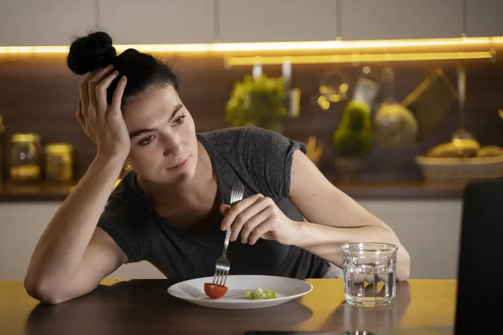 A woman sitting in her kitchen, looking frustrated while eating a small portion of salad, reflecting on the idea that exercise might be keeping people fat and the unspoken challenges of weight loss. Exercise Is Keeping People Fat But No One Wants to Talk About It