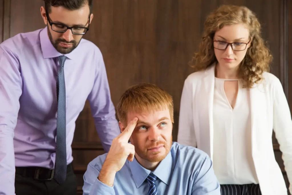 Three people in formal attire are gathered around a table; a man in the center appears stressed, pondering perhaps the irony that exercise might be keeping people fat, while the other two scrutinize documents, seemingly oblivious to this unspoken dilemma.